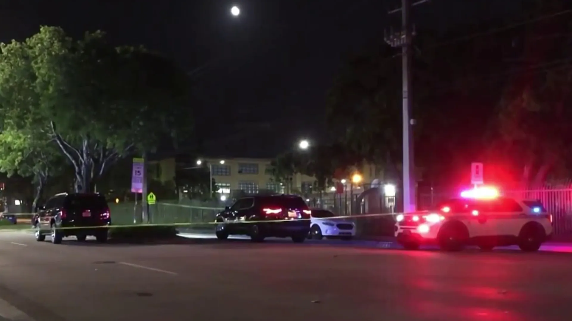 Marcadores de evidencia en el suelo tras un tiroteo en La Pequeña Haití, Miami, con vehículos policiales al fondo.