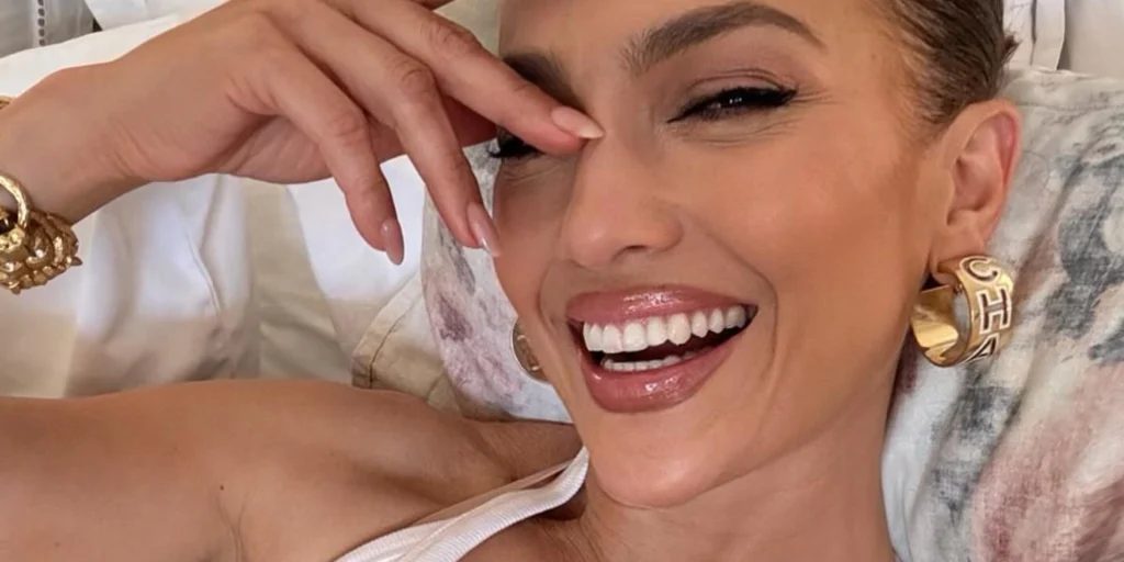 Jennifer Lopez sonriendo en su fiesta de cumpleaños con un hombre de expresión seria al fondo.