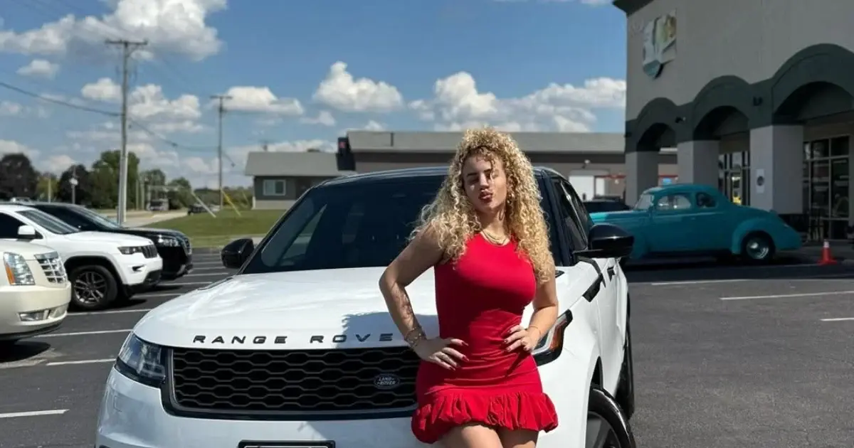 Fotografía conceptual y realista de Flor de Cuba sonriendo junto a un moderno Range Rover blanco, mientras en un segundo plano se vislumbra un Toyota Corolla siendo cargado para envío. Estilo cinematográfico, alta definición.
